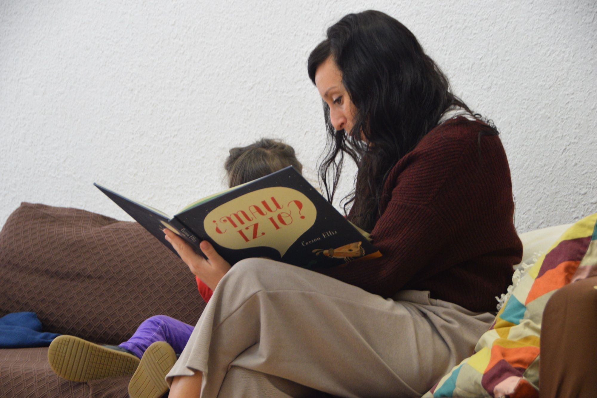 A teacher reads a picture book on the sofa, with a child beside her